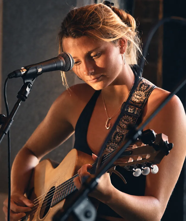 Lucy Spotts singing into a microphone while holding an acoustic guitar.